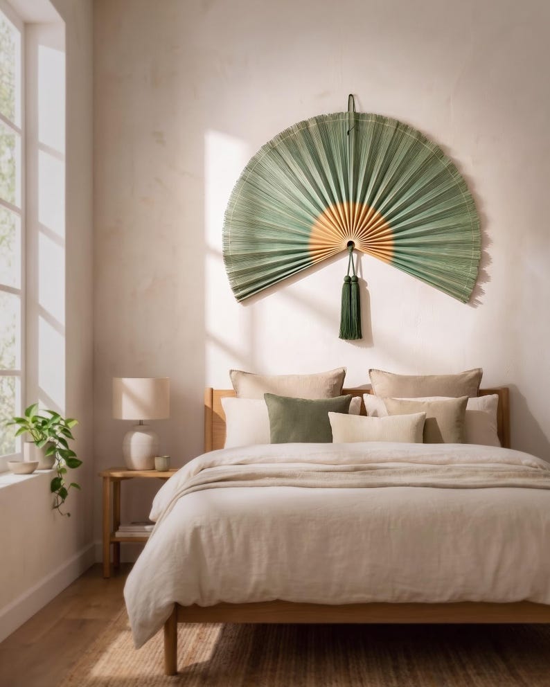 May include: A large, semi-circular wall fan made of pleated green palm leaves with a natural light wood center. The fan features a long, dark green decorative tassel hanging from the bottom center and is mounted on a wall above a bed.