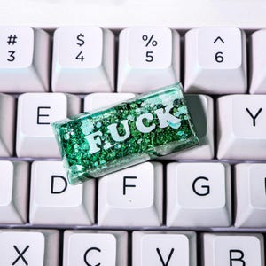 May include: A rectangular, clear resin object filled with green glitter and the word "FUCK" in white letters, resting on a white keyboard. The keyboard keys are arranged in a standard QWERTY layout.