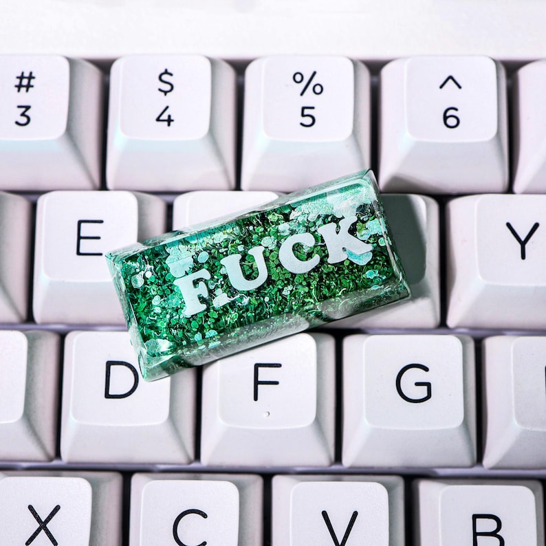May include: A rectangular, clear resin object filled with green glitter and the word "FUCK" in white letters, resting on a white keyboard. The keyboard keys are arranged in a standard QWERTY layout.