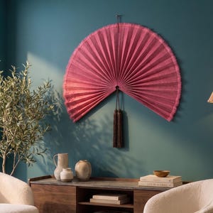 May include: A large, semi-circular wall fan in a deep magenta pink hue with a pleated texture and a fringed edge. A long, dark brown decorative tassel hangs from the center point of the fan, which is mounted securely against a solid wall surface.