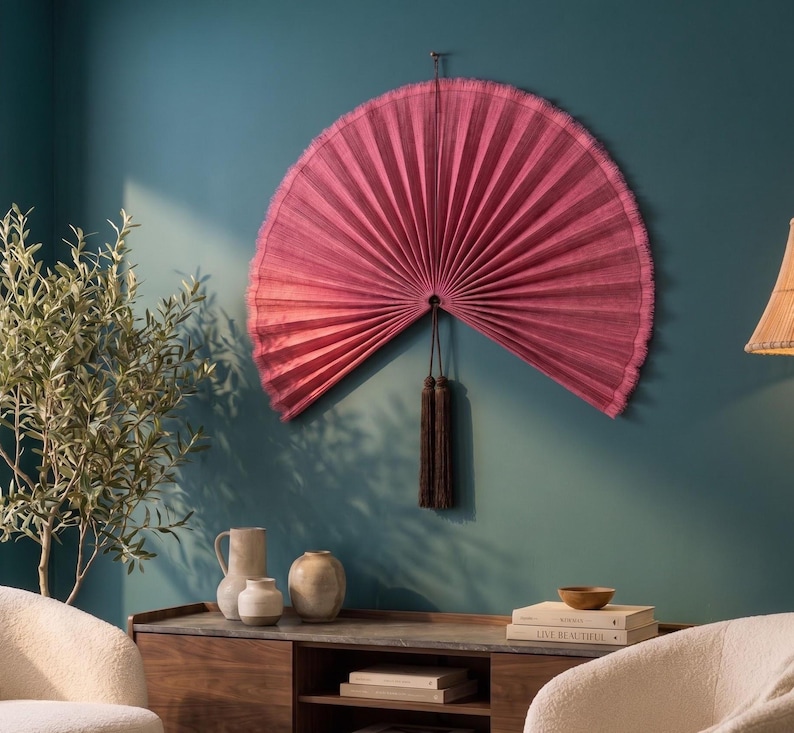 May include: A large, semi-circular wall fan in a deep magenta pink hue with a pleated texture and a fringed edge. A long, dark brown decorative tassel hangs from the center point of the fan, which is mounted securely against a solid wall surface.