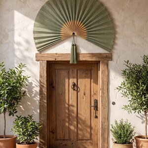 May include: A large, semi-circular wall fan made of pleated, sage green palm leaves with a natural tan center. The fan features a decorative sage green tassel hanging from the bottom center, positioned above a wooden door with a metal door knocker.