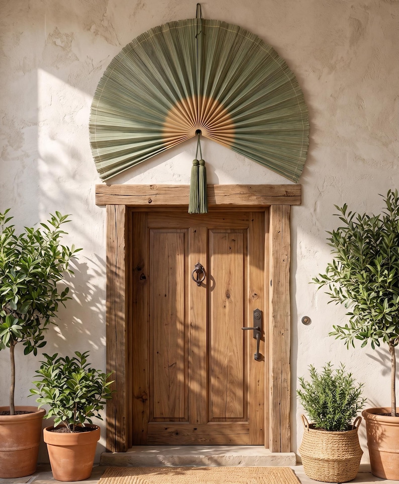 May include: A large, semi-circular wall fan made of pleated, sage green palm leaves with a natural tan center. The fan features a decorative sage green tassel hanging from the bottom center, positioned above a wooden door with a metal door knocker.