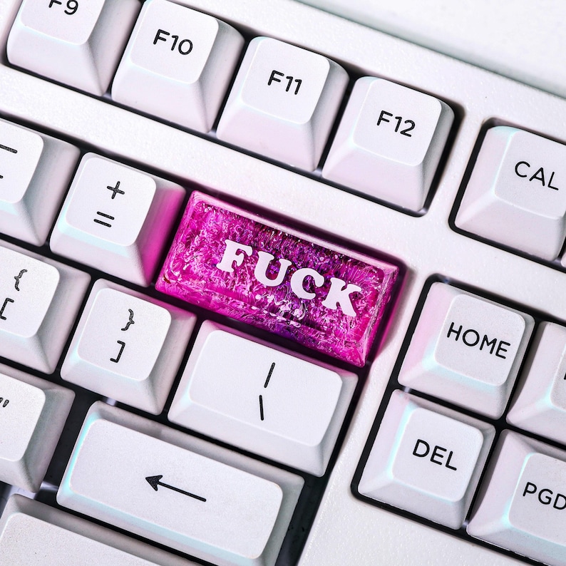 May include: Close-up of a white computer keyboard with a custom pink key that reads "FUCK" in white capital letters. The key is surrounded by other white keys with function keys and symbols.