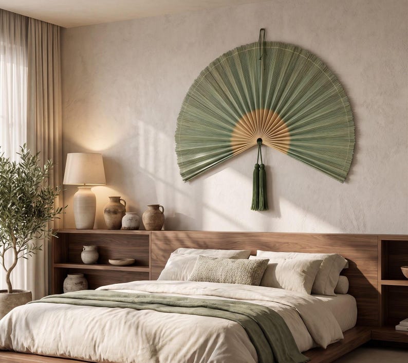 May include: A large, semi-circular wall fan made of pleated green and tan natural fibers, featuring a central pivot point and a long, dark green decorative tassel hanging from the bottom. The fan is mounted on a wall above a wooden headboard.