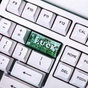 May include: Close-up of a white computer keyboard with a custom keycap. The keycap is green with glitter and the word "FUCK" in white. Other keys include F9, F10, F11, F12, DEL, HOME, and CAL.