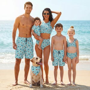 May include: A family of six, including a dog, is on a beach. They are wearing matching light blue swimwear with a pattern of red starfish and blue floral designs. The dog wears a matching bandana.