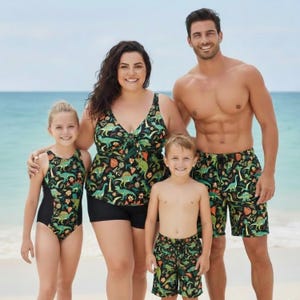 May include: A family of four on a beach, wearing matching dinosaur-themed swimwear. The swimwear includes a one-piece swimsuit, tankini top with shorts, and swim shorts. The color scheme is black with green and orange dinosaur illustrations.
