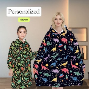 Pode incluir: Duas mantas com capuz vestíveis. Uma é preta com dinossauros coloridos, planetas e naves espaciais. A outra é verde com várias ilustrações de dinossauros. O texto "Personalized PHOTO" está no topo.