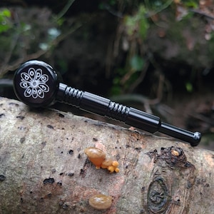 May include: Black metal pipe with a white floral design on the bowl. The pipe is on a tree branch with small, tan fungi. The background is blurred, showing a natural outdoor setting.