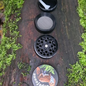 May include: A black, four-piece herb grinder with a squirrel illustration on the bottom piece. The grinder is disassembled, showing the top lid with a white scraper, a mesh screen, and the grinding chamber. The bottom piece has the text "dopefauna".