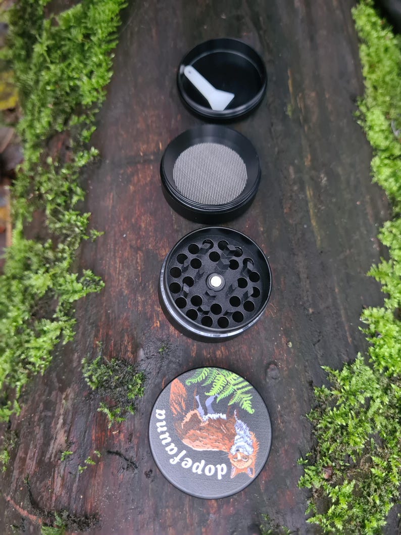 May include: A black, four-piece herb grinder with a squirrel illustration on the bottom piece. The grinder is disassembled, showing the top lid with a white scraper, a mesh screen, and the grinding chamber. The bottom piece has the text "dopefauna".