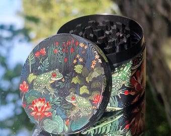 May include: A black and green herb grinder with a detailed frog and lily pad design. The lid features two green frogs with red eyes, surrounded by red flowers and foliage. The cylindrical body has a matching pattern.