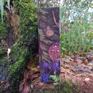May include: A rectangular product package with a nature-themed design. The package features illustrations of a monarch butterfly, a mushroom, and blue thistle flowers against a camouflage background. The package is set against a natural, mossy backdrop.
