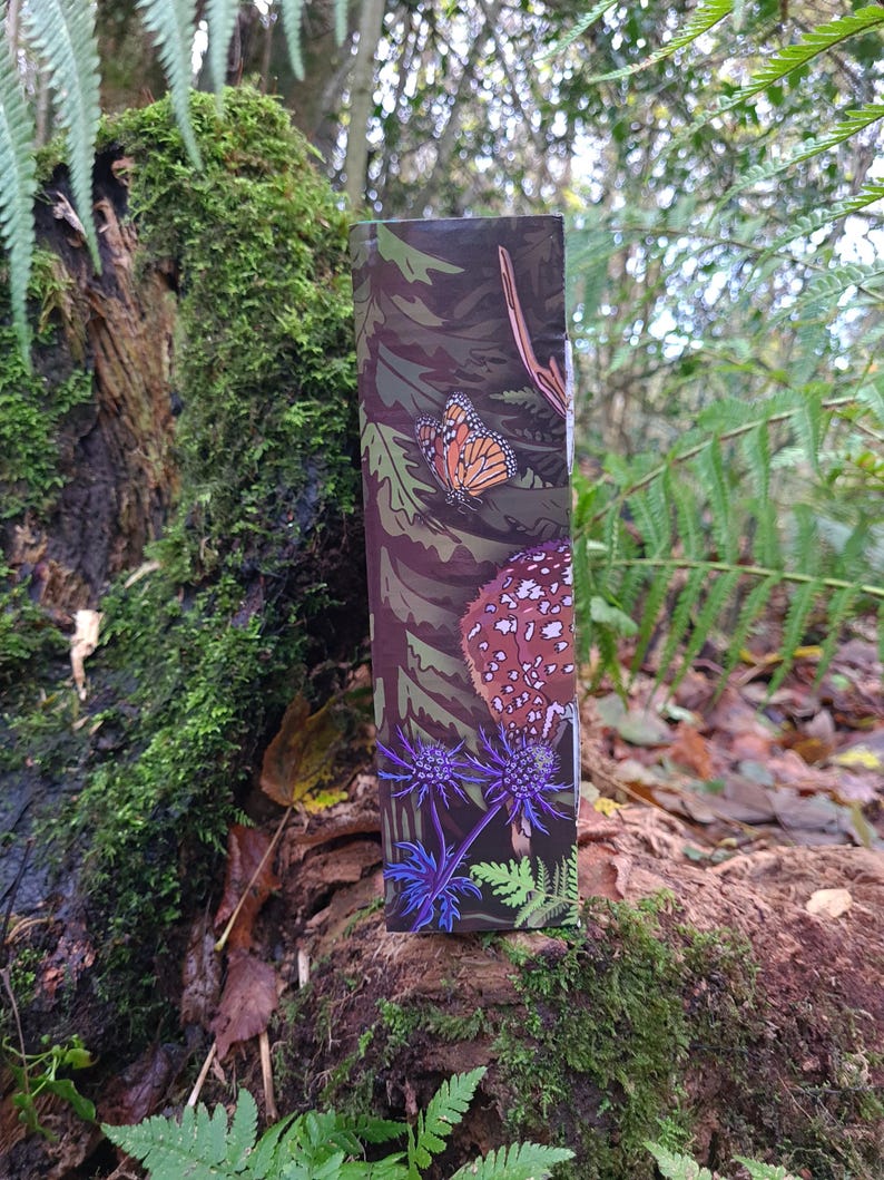 May include: A rectangular product package with a nature-themed design. The package features illustrations of a monarch butterfly, a mushroom, and blue thistle flowers against a camouflage background. The package is set against a natural, mossy backdrop.