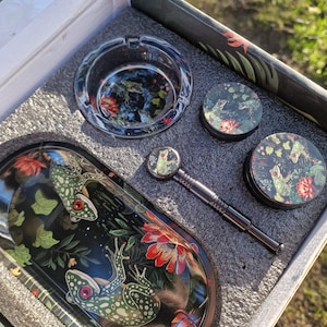Op de afbeelding: Een glazen asbak met een kikker- en bloemenontwerp, een klein glazen potje met een kikker- en bloemenontwerp, een metalen pokertool en een glazen dienblad met een kikker- en bloemenontwerp.