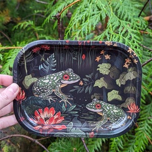 Pode incluir: Bandeja retangular preta com uma ilustração detalhada de duas rãs verdes com olhos vermelhos, rodeadas por flores vermelhas e nenúfares. A bandeja tem um fundo escuro com um tema de floresta.