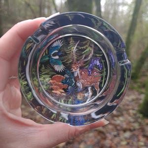 May include: A clear glass ashtray with a detailed forest scene featuring a deer, bird, and flowers. The ashtray has a dark rim and is held against a blurred forest background.