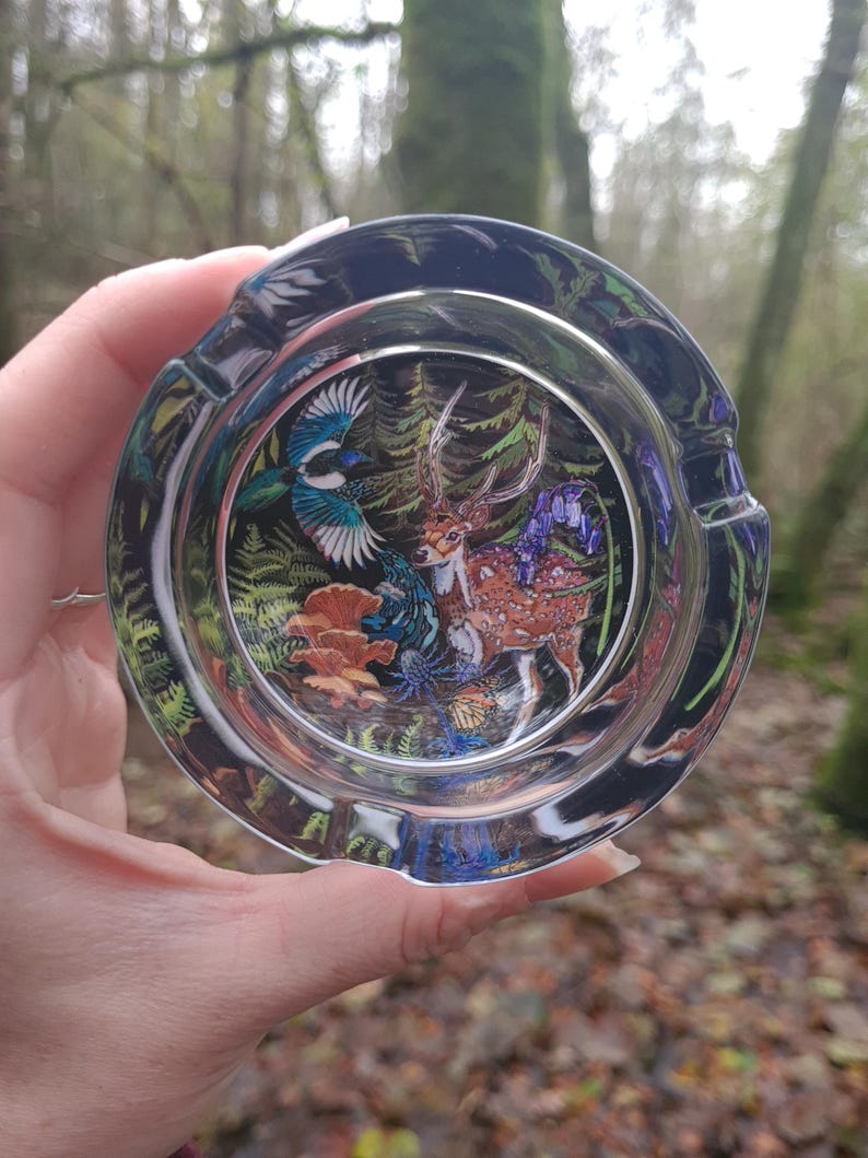 May include: A clear glass ashtray with a detailed forest scene featuring a deer, bird, and flowers. The ashtray has a dark rim and is held against a blurred forest background.