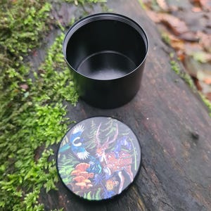 May include: A black, cylindrical container with a lid featuring a colorful illustration of a deer, bird, and mushrooms. The container is open, revealing its interior. The lid is placed next to the container on a wooden surface.
