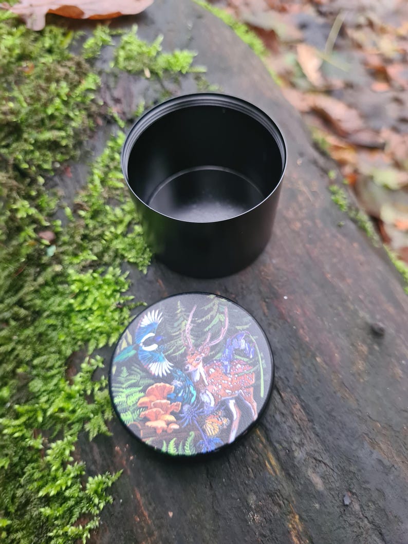 May include: A black, cylindrical container with a lid featuring a colorful illustration of a deer, bird, and mushrooms. The container is open, revealing its interior. The lid is placed next to the container on a wooden surface.