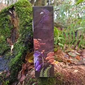May include: A rectangular box with a dark brown background featuring a purple raven illustration. The box also displays illustrations of purple flowers, orange mushrooms, and green ferns. The box is set against a natural forest backdrop.
