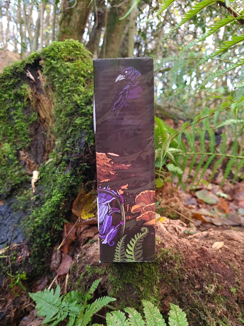 May include: A rectangular box with a dark brown background featuring a purple raven illustration. The box also displays illustrations of purple flowers, orange mushrooms, and green ferns. The box is set against a natural forest backdrop.