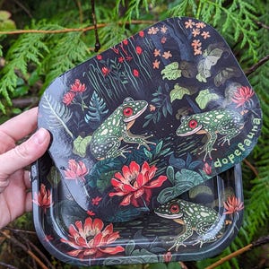 Pode incluir: Uma bandeja de metal preta com um design floral verde e vermelho com sapos e o texto "dopefauna".