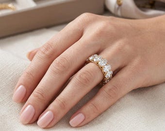 May include: A gold band ring set with multiple oval-cut diamonds. The ring is on a person's finger, with the hand resting on a light-colored surface. The ring's diamonds sparkle, and the gold band provides a warm contrast.