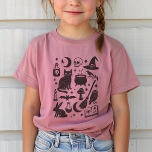 May include: A mauve-colored t-shirt featuring a black graphic of Halloween-themed icons. The design includes a cat, skull, witch's hat, cauldron, and other spooky elements. The shirt is worn by a young person.
