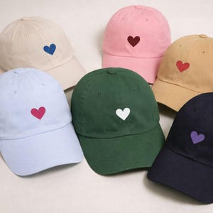 Peut inclure: Plusieurs casquettes de baseball de couleurs variées, dont bleu clair, rose, beige, vert et marine. Chaque casquette est ornée d'un motif brodé en forme de cœur de couleur contrastante. Les casquettes sont en tissu doux et ont une visière incurvée.