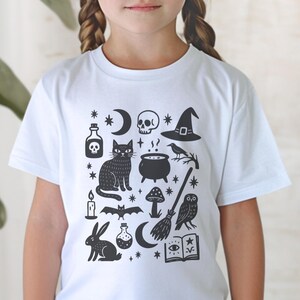 May include: White t-shirt featuring a black graphic design with witchy and Halloween-themed illustrations. The design includes a black cat, skull, cauldron, moon, stars, witch hat, owl, and other mystical elements.