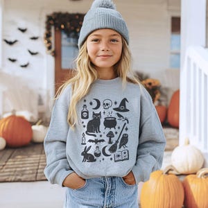 May include: A light grey sweatshirt featuring a black Halloween graphic with a cat, skull, witch's hat, and cauldron. The person is wearing a grey beanie and blue jeans. Pumpkins are in the background.