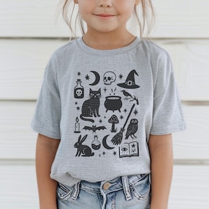 May include: A gray t-shirt featuring a black graphic of Halloween-themed icons. The design includes a skull, witch hat, black cat, cauldron, owl, and other spooky elements. The shirt is a short-sleeved crew neck.