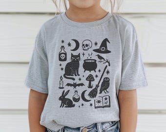 May include: A gray t-shirt featuring a black graphic of Halloween-themed icons. The design includes a skull, witch hat, black cat, cauldron, owl, and other spooky elements. The shirt is a short-sleeved crew neck.