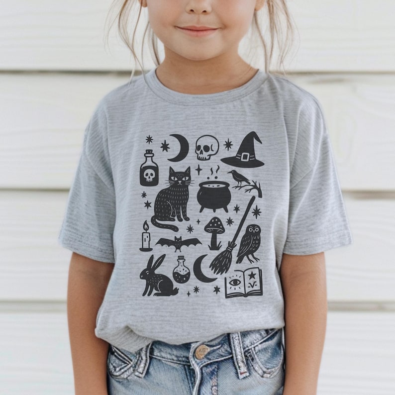 May include: A gray t-shirt featuring a black graphic of Halloween-themed icons. The design includes a skull, witch hat, black cat, cauldron, owl, and other spooky elements. The shirt is a short-sleeved crew neck.