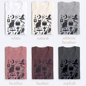 May include: Six t-shirts in various colors, each featuring a black graphic design with witchy elements. The design includes a cat, cauldron, moon, skull, witch hat, and other mystical symbols. Colors include white, natural, athletic heather, heather mauve, heather maroon, and asphalt.