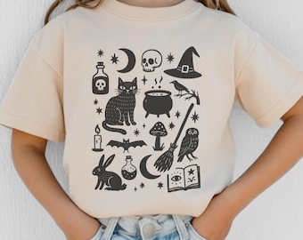 May include: Beige short-sleeved t-shirt featuring a black graphic design of Halloween-themed illustrations. The design includes a cat, skull, witch hat, potion bottles, a cauldron, a broom, and other spooky elements.