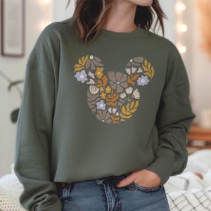 Pu&ograve; includere: Una felpa verde con un design floreale di Topolino. I fiori sono in tonalit&agrave; di giallo, arancione, rosa e viola.