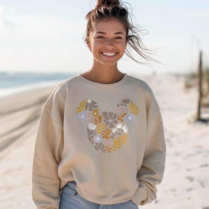 Pu&ograve; includere: Una felpa beige con un design floreale di Topolino. I fiori sono in tonalit&agrave; di giallo, arancione, rosa e viola.