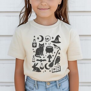 May include: A cream-colored t-shirt featuring a black graphic design of Halloween-themed icons. The design includes a cat, skull, witch hat, potion bottles, a cauldron, a broom, and other spooky elements. The shirt is made of soft material.