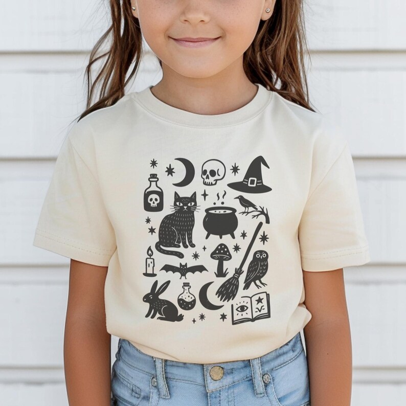May include: A cream-colored t-shirt featuring a black graphic design of Halloween-themed icons. The design includes a cat, skull, witch hat, potion bottles, a cauldron, a broom, and other spooky elements. The shirt is made of soft material.