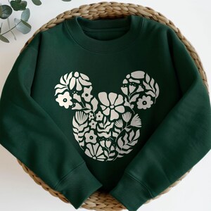 Pu&ograve; includere: Una felpa verde scuro con un design floreale di Topolino bianco.