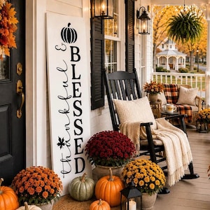 May include: A tall white vertical wooden porch sign featuring the black text Thankful & Blessed with a pumpkin icon at the top and a maple leaf icon at the bottom. The sign is displayed on a porch next to a black rocking chair and pumpkins.