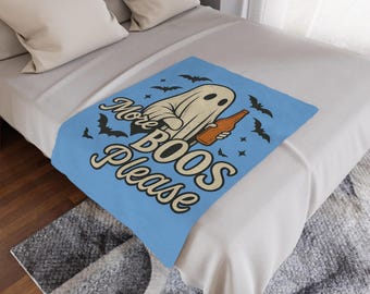 May include: A light blue blanket with a Halloween theme. The blanket features a cartoon ghost holding a bottle, with the text "More BOOS Please" and black bat illustrations. The blanket is on a bed with white pillows.