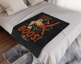 May include: Black throw blanket with a Halloween-themed graphic. The design features a ghost, a cat, and the text "I'M HERE FOR THE BOOS!" in orange and white. The blanket is displayed on a bed with white and gray bedding.