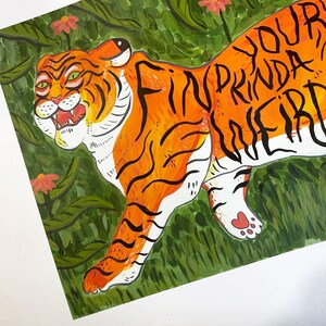 May include: A watercolor painting of an orange and black tiger with green foliage and pink flowers in the background. The tiger has the words "Find your kinda weird" written on its body.