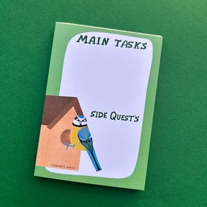 May include: A notepad with a green border and a white interior, featuring the text "MAIN TASKS" and "SIDE QUEST'S". A colourful illustration of a blue and yellow bird perched near a birdhouse is on the left side. The notepad is on a green surface.