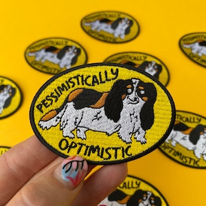 May include: Yellow embroidered iron-on patch featuring a cartoon Cavalier King Charles Spaniel dog. The dog is black, brown, and white. The text on the patch reads "Pessimistically Optimistic".