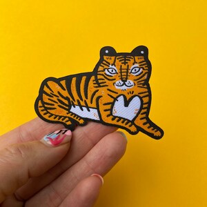 May include: An iron-on patch featuring an embroidered tiger lying down. The tiger is yellow and black with white stripes and a white belly.